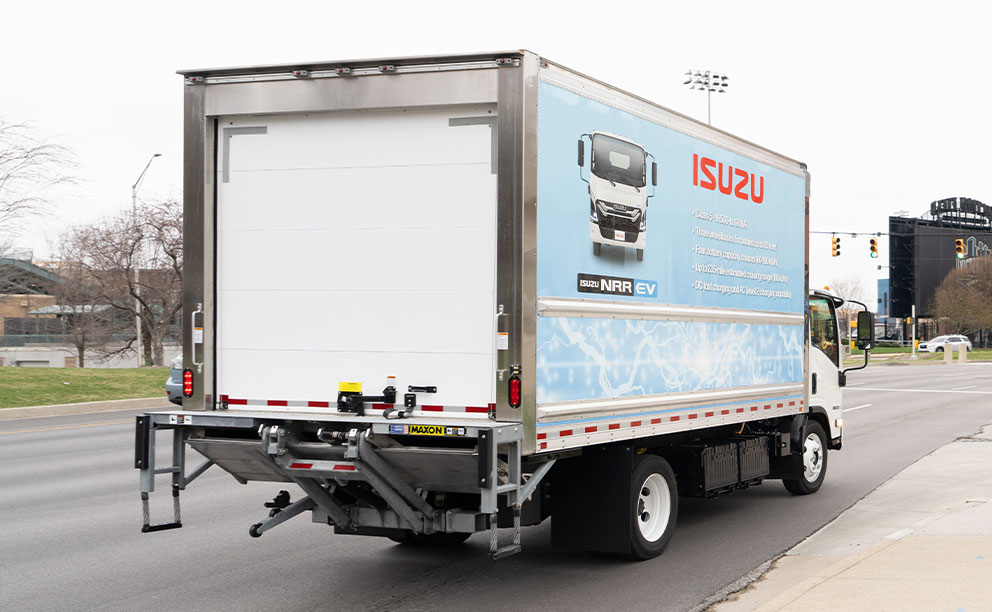 Small, white dry freight body with blue decals, mounted on Isuzu NPR electric chassis, photographed from behind while driving on a highway.
